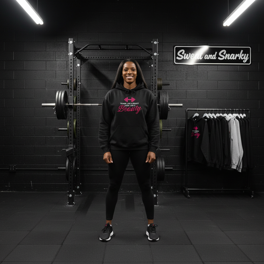 Woman in a gym wearing a black hoodie with pink text, standing in front of weightlifting equipment.